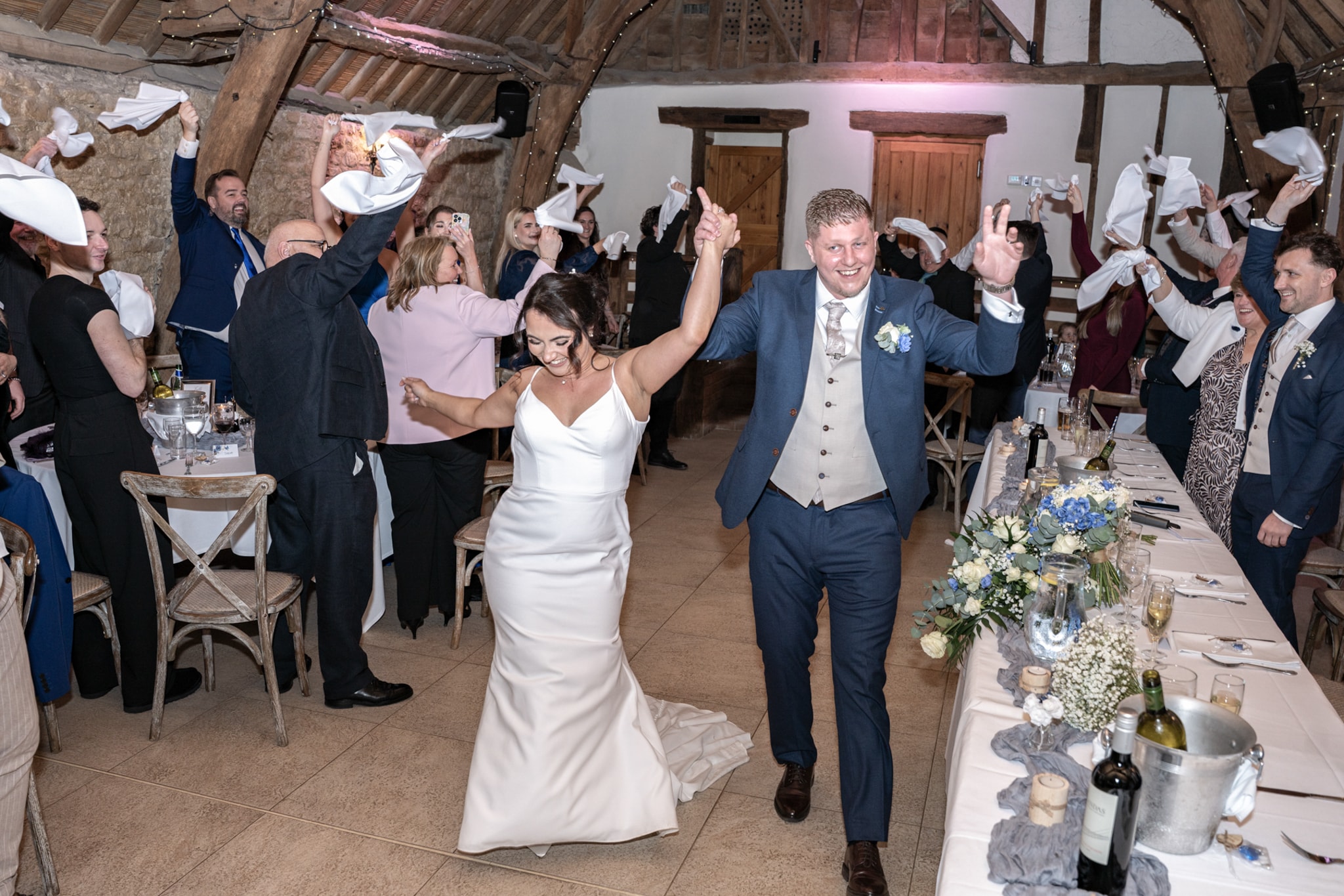 Bride and groom entering reception room with guests waving napkins in celebration at Notley Tythe Barn