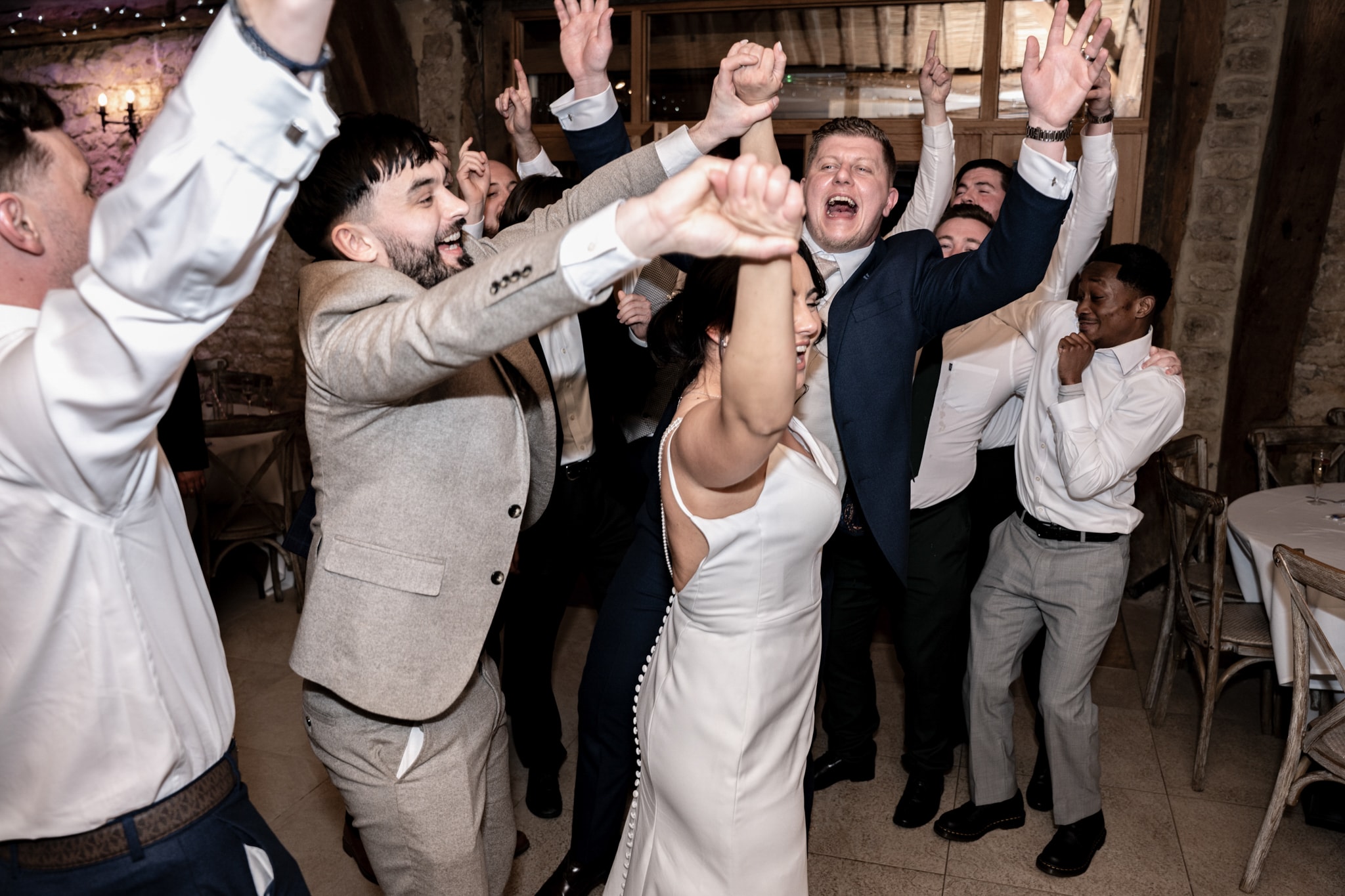 Bride and wedding guests celebrating with arms raised on dance floor at Notley Tythe Barn reception in colour