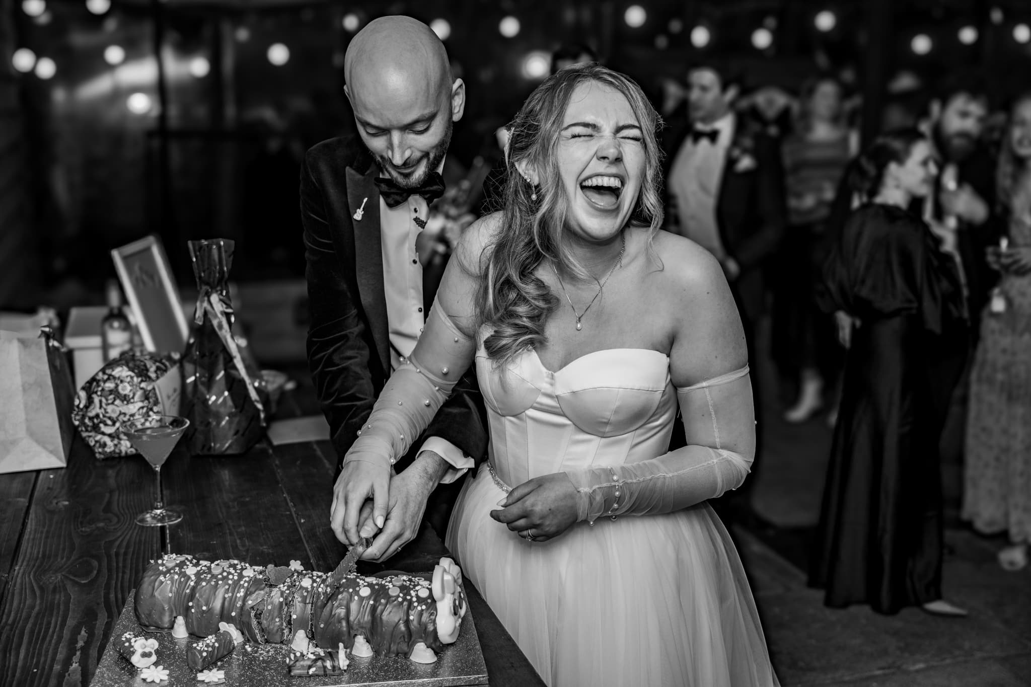 bride laughing whilst cutting wedding cake black and white image